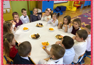 Dzieci elegancko ubrane siedzą przy stole nakrytym białym obrusem i częstują się słodyczami.